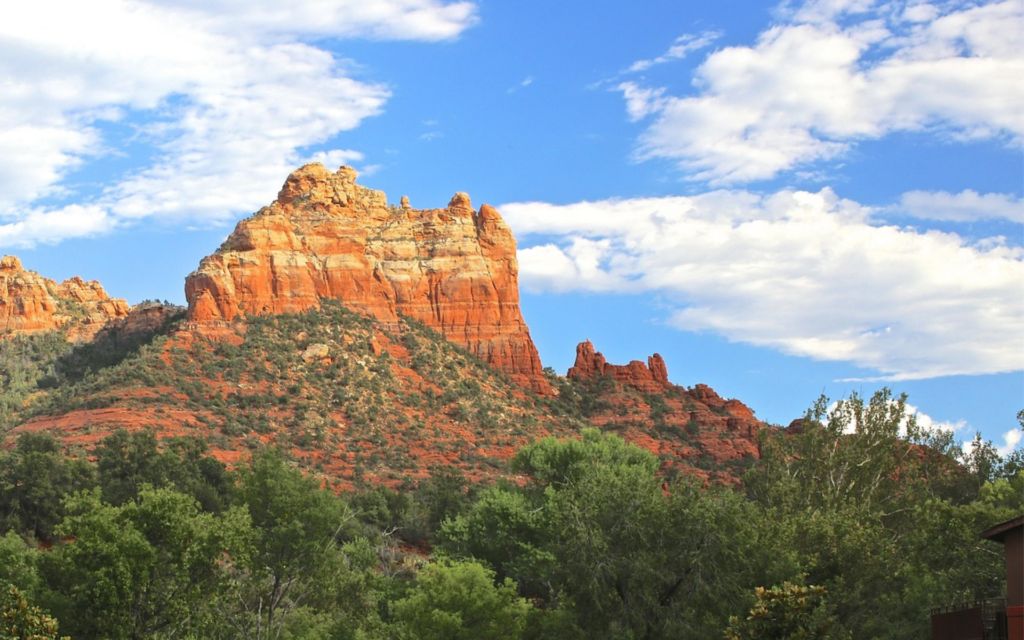 Signature red rocks of Uptown sedona with lush green forest at the base of the mountain with blue, cloudy skies.
